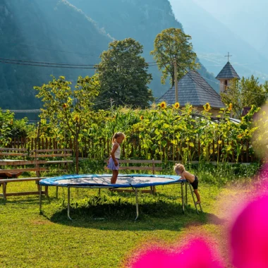Twee kinderen spelen bij een trampoline in de tuin van een guesthouse in Theth, Albanië, met zonnebloemen en bergen op de achtergrond