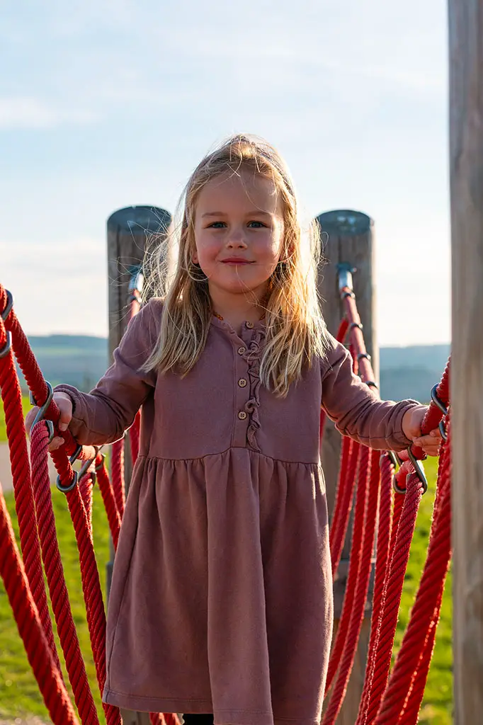 Child playing on a small red rope climbing frame on the way to the Geierlay Suspension Bridge in Germany, with a hilly landscape and blue sky in the background.