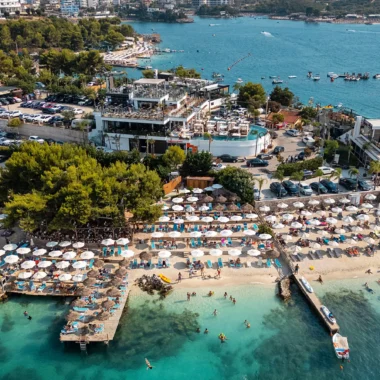 Luchtfoto van een druk strand in Ksamil, met turquoise water, rijen parasols, strandbedjes en een groot beachclubcomplex tijdens een vakantie in Albanië. Op de achtergrond zijn heuvels, boten en een levendige kustlijn zichtbaar.