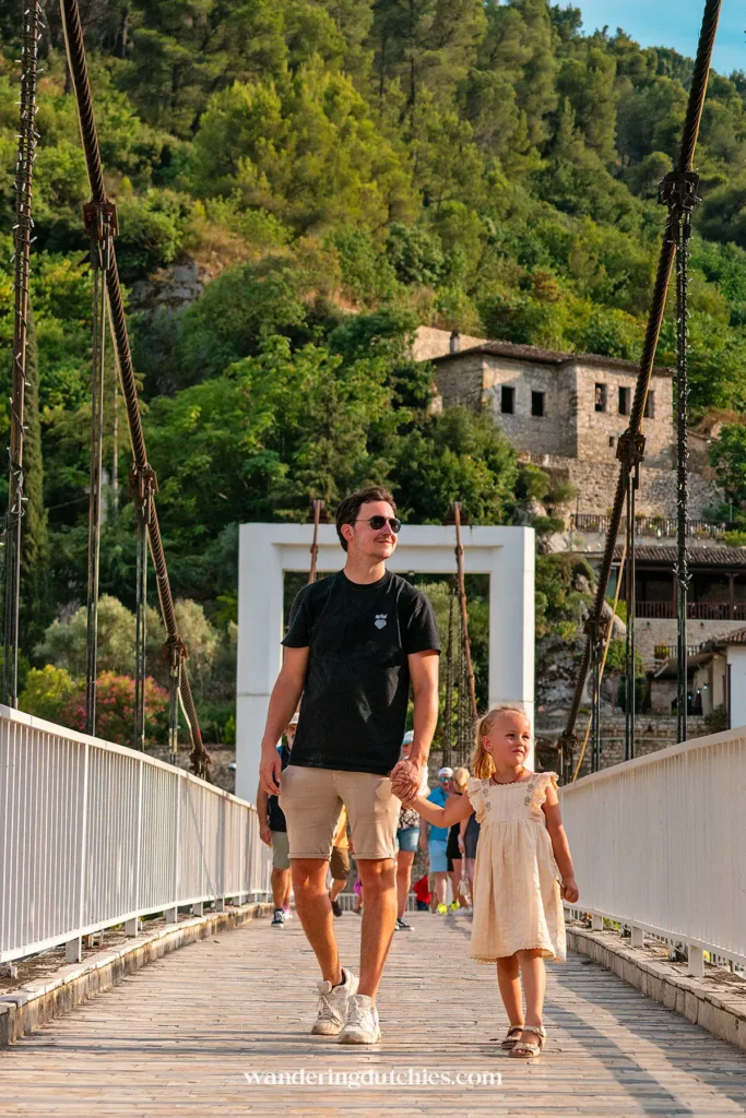 Vader en dochter wandelen over een brug met uitzicht op de groene heuvels en stenen gebouwen van Berat, Albanië.