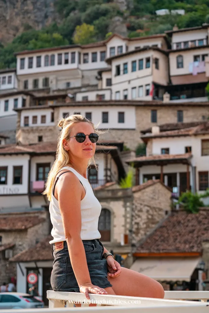 Portret van een vrouw met zonnebril, zittend voor de traditionele huizen met witte gevels en rode daken van Berat, Albanië.