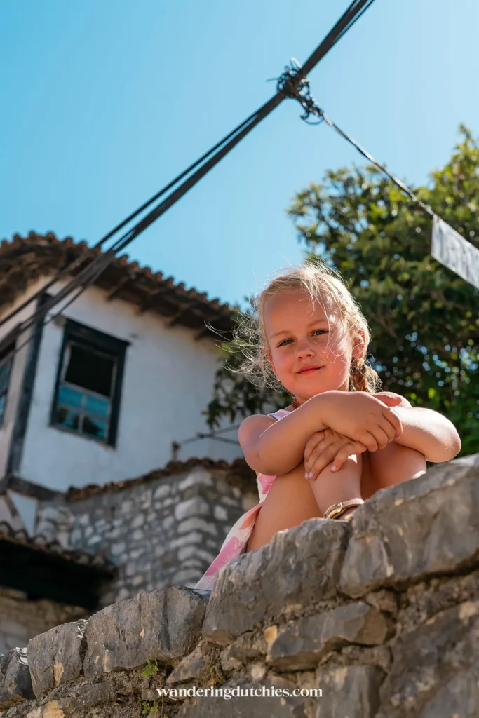 Een jong meisje zittend op een muur in Berat Castle, met een traditionele woning op de achtergrond.