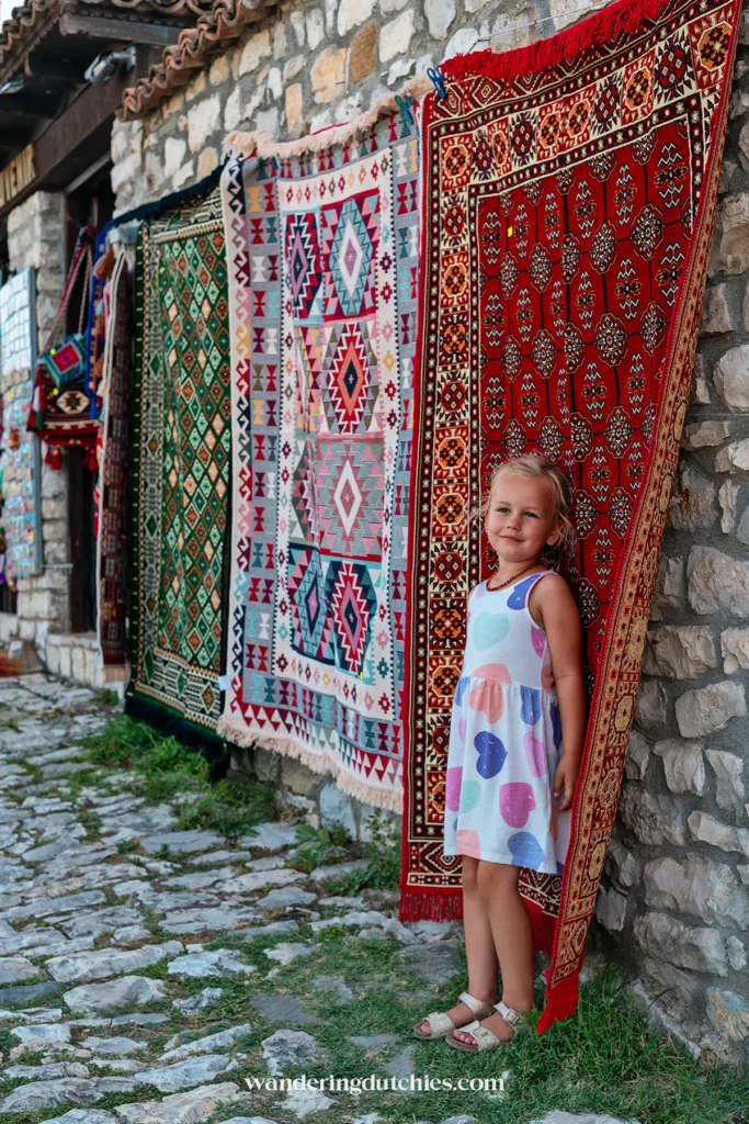 Een jong meisje staat naast kleurrijke handgeweven tapijten in Berat Castle.