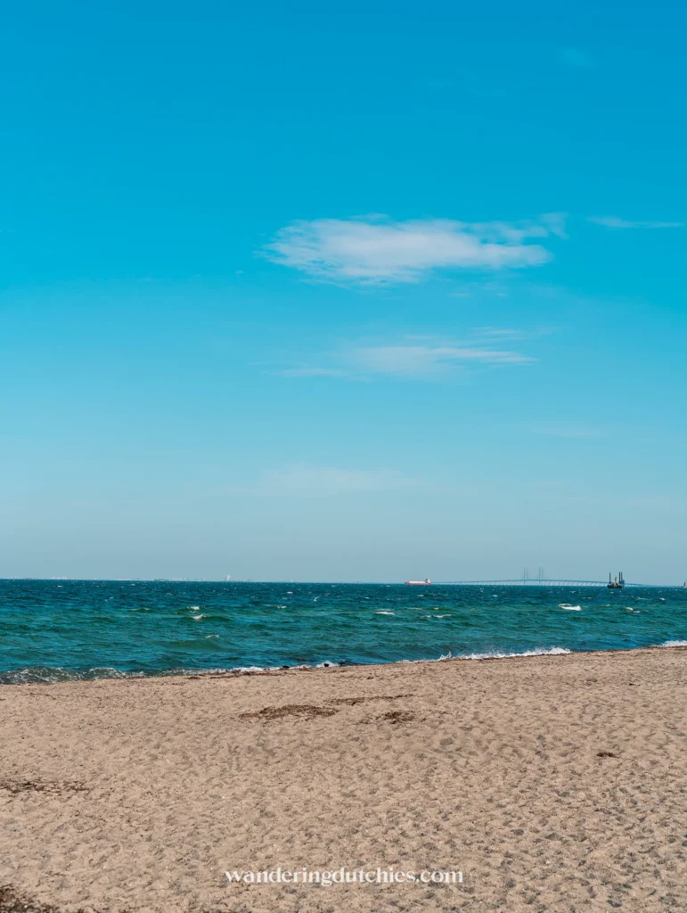 Uitzicht op het Amager Strand in Kopenhagen met zand, de Øresundbrug in de verte en een helderblauwe lucht boven het water.