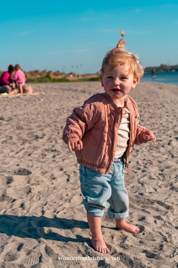 Een jong kind loopt en speelt op het zand van Amager Strand in Kopenhagen, met de zee en andere bezoekers op de achtergrond.