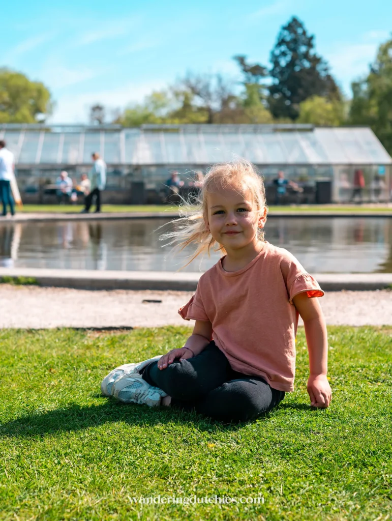 Een jong kind zit op het gras voor een grote kas in Botanisk Have, Kopenhagen.