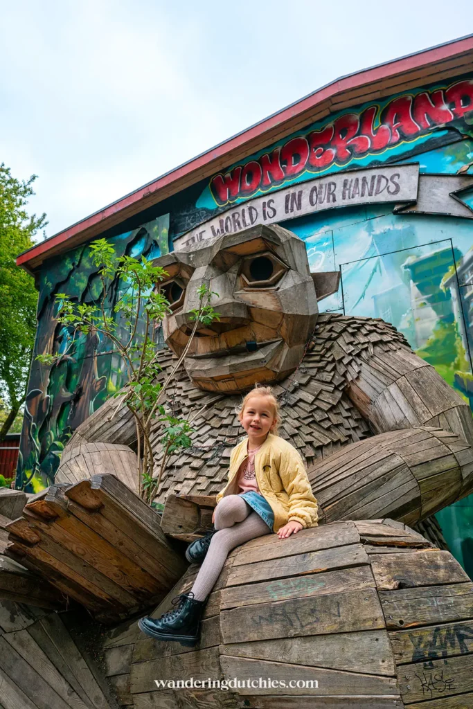 Child sitting on a large wooden troll by Thomas Dambo in Christiania, Copenhagen, surrounded by colorful street art.