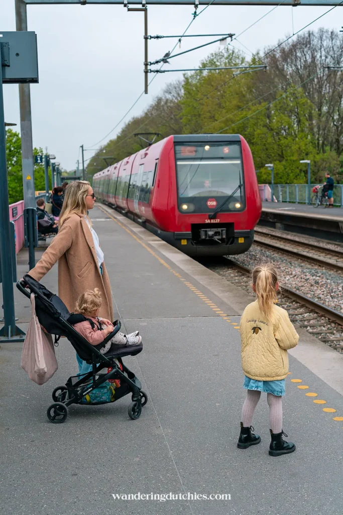 Moeder met kinderwagen en dochter wachten op het perron terwijl een rode S-trein arriveert in Kopenhagen.