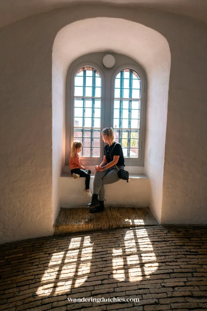 Moeder en kind zitten bij een groot raam in de Rundetaarn, met licht dat door de ramen valt.