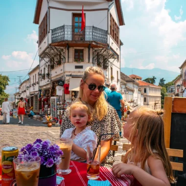 Mother and two young children enjoying drinks at a terrace in Gjirokastër, Albania, with a traditional building in the background.