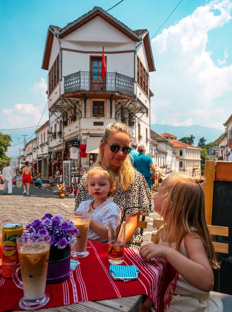 Mother and two young children enjoying drinks at a terrace in Gjirokastër, Albania, with a traditional building in the background.