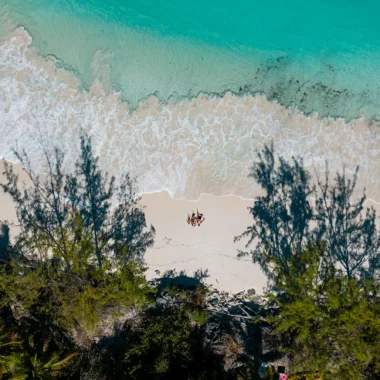 Luchtfoto van een tropisch strand op Exuma, Bahama’s, met helder turquoise water dat zachtjes over het witte zand spoelt. In het midden ligt een gezin ontspannen op het strand, omringd door schaduwen van palmbomen.
