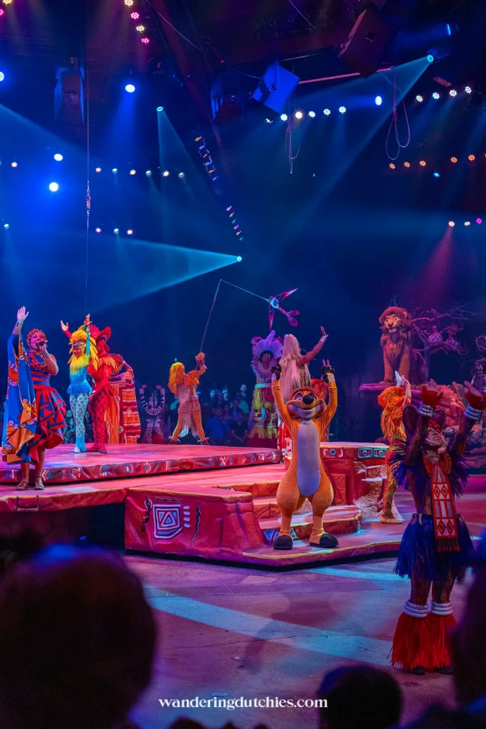 Dynamische scène van de Festival of the Lion King show met artiesten in kleurrijke kostuums en bekende Disney-figuren.
