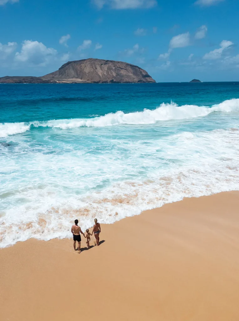 Gezin wandelt langs het strand van Playa de las Conchas op La Graciosa, met uitzicht op het eiland Montaña Clara in de verte en helderblauw zeewater op de achtergrond.