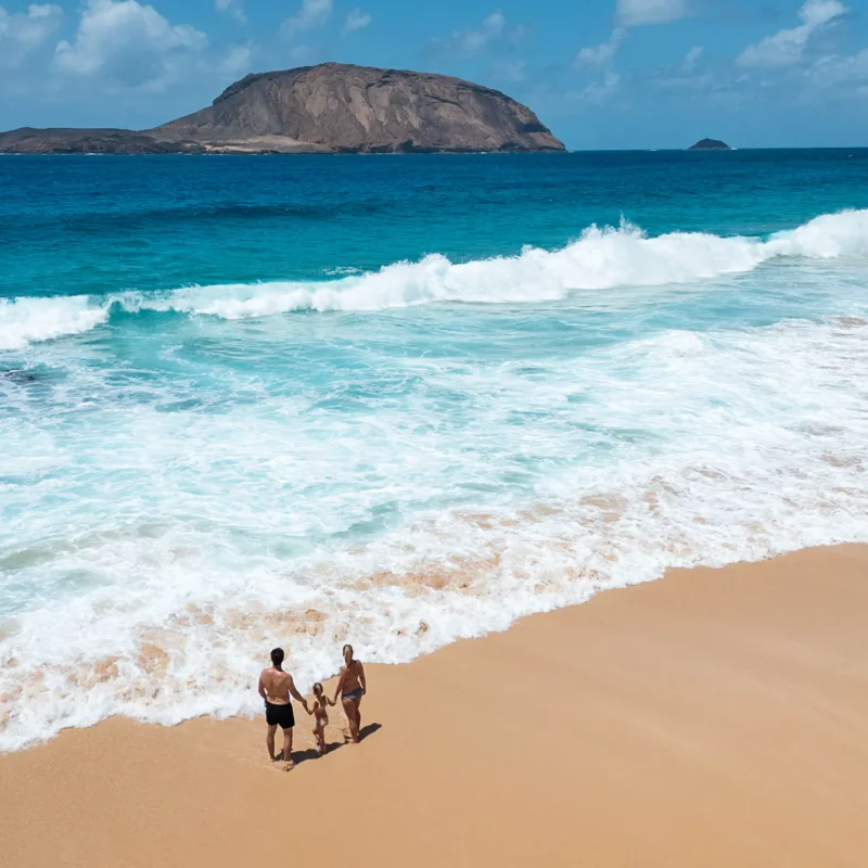 Gezin wandelt langs het strand van Playa de las Conchas op La Graciosa, met uitzicht op het eiland Montaña Clara in de verte en helderblauw zeewater op de achtergrond.