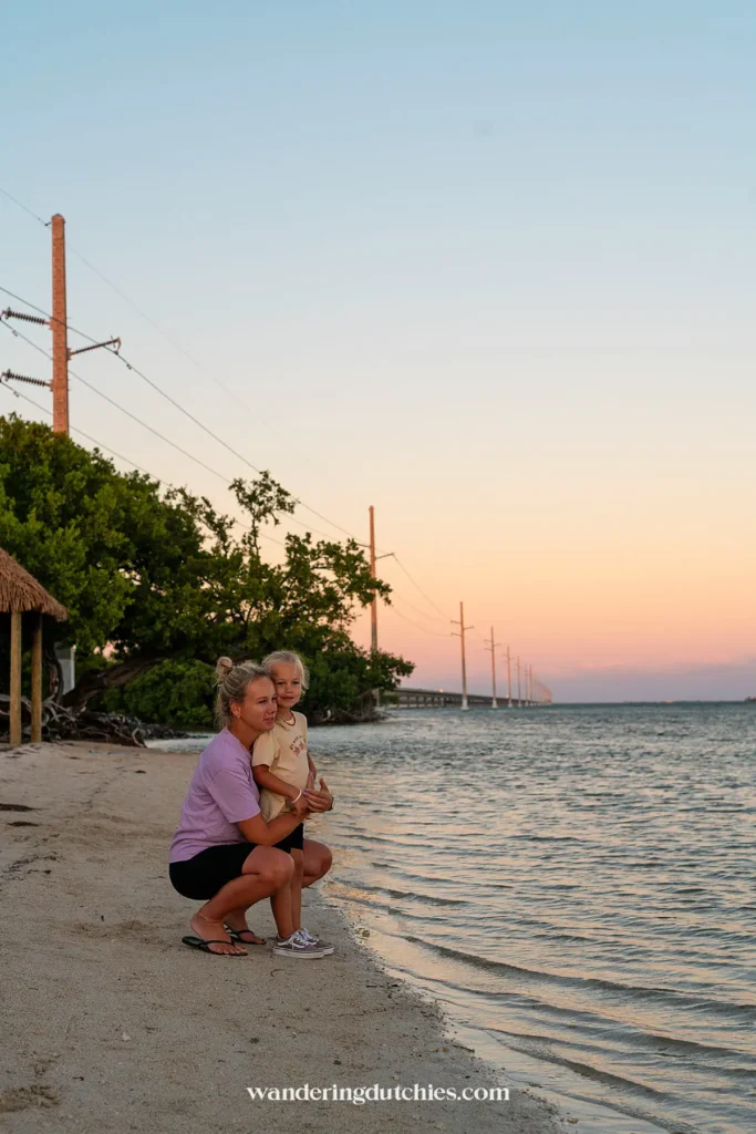 Moeder knielt met dochter aan de waterkant bij zonsondergang, rustig strand met palen en de Seven Mile Bridge op de achtergrond in de Florida Keys