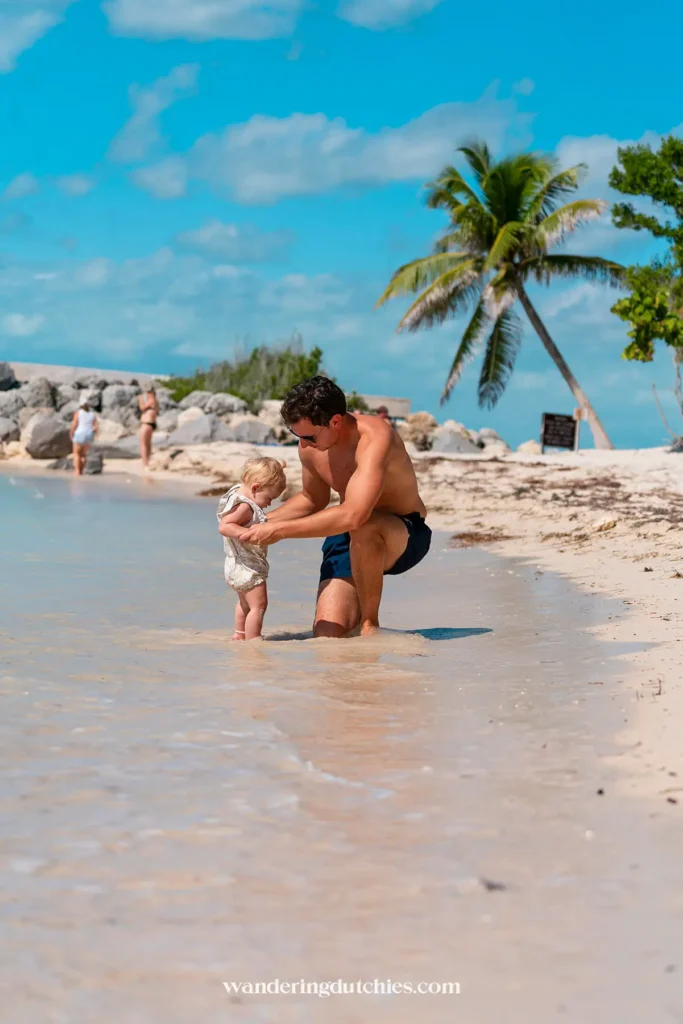 Vader helpt peuter pootjebaden op een ondiep strand met palmboom op de achtergrond in de Florida Keys.