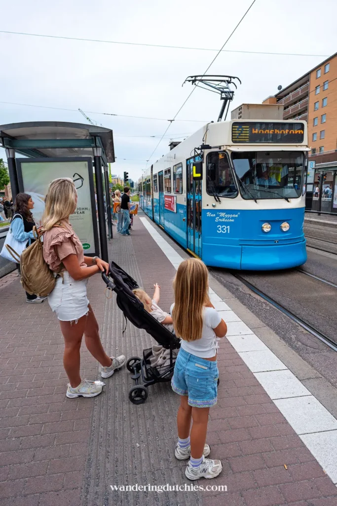 Vrouw met kinderwagen en twee kinderen wacht op de blauwe tram in Göteborg.