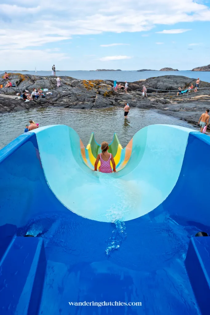 Kind glijdt van blauwe waterglijbaan het natuurlijke zeebad in bij Åstol, Zweden.