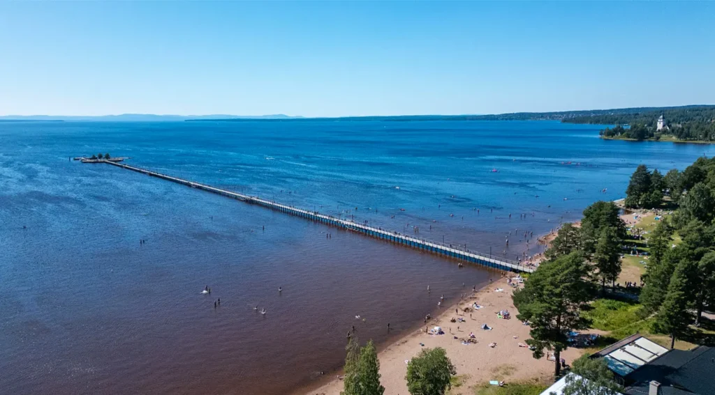 Long wooden pier stretching into Lake Siljan in Rättvik, with beach and swimmers along the shore.