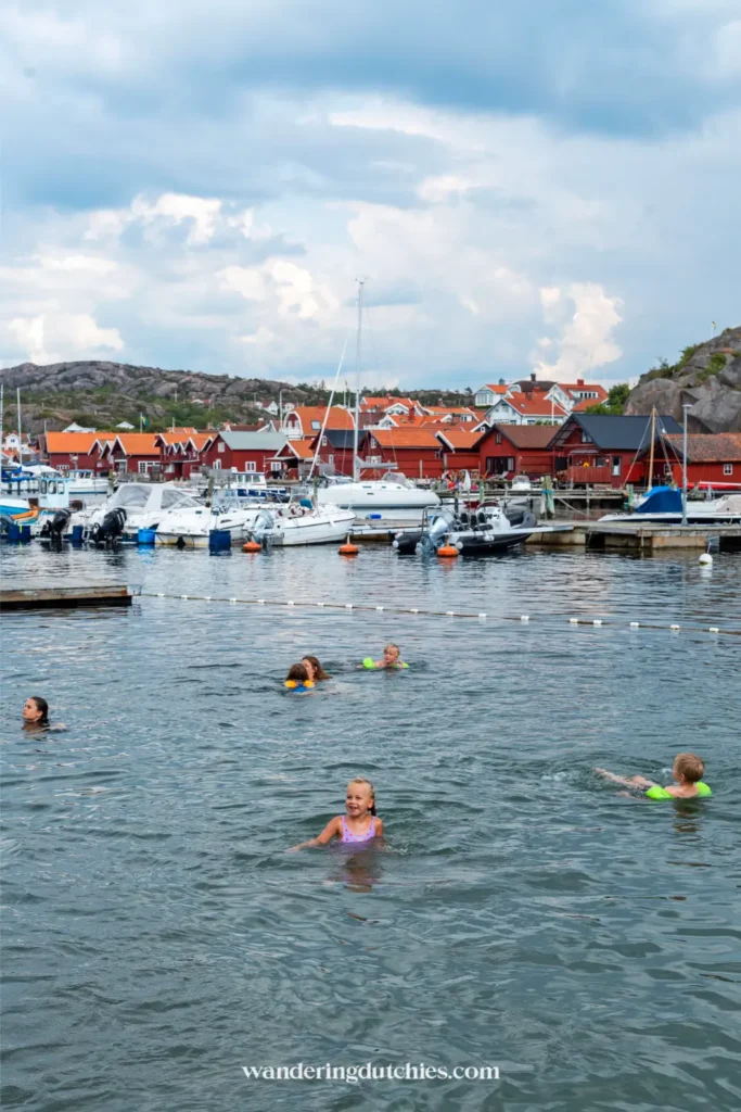 Kinderen zwemmen in het water van de haven van Bovallstrand met boten en rode houten huizen op de achtergrond