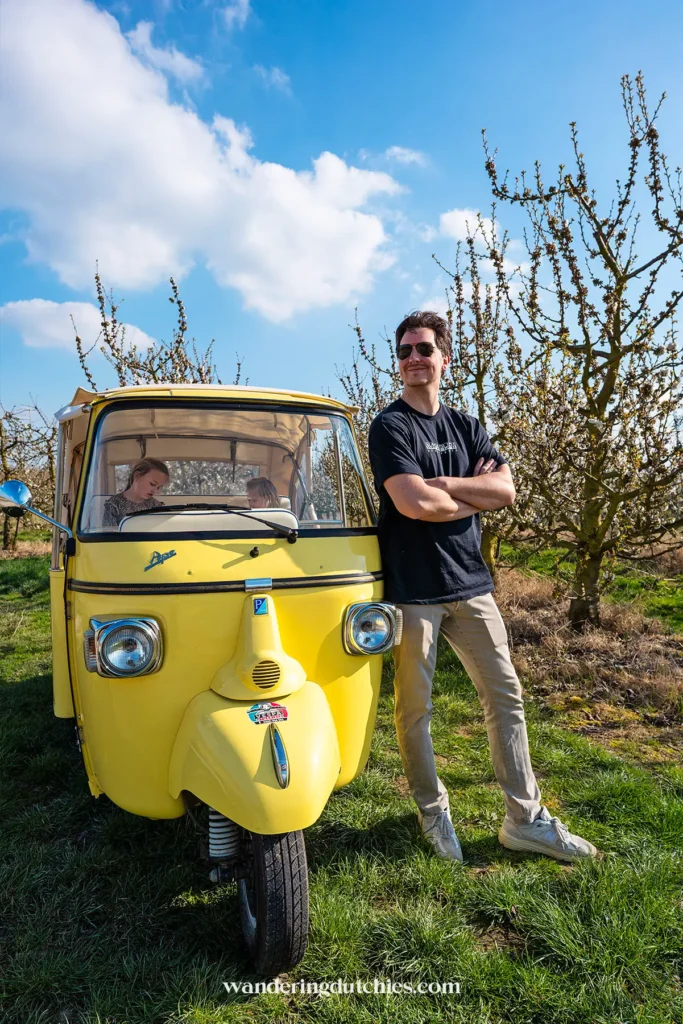 Man bij gele Ape in bloesemboomgaard tijdens roadtrip in Borgloon, België.