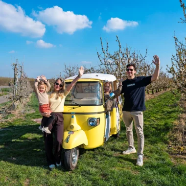 Gezin bij een gele Ape Calessino tijdens de Bambinotour tussen fruitboomgaarden in Tongeren-Borgloon, Belgisch Limburg.