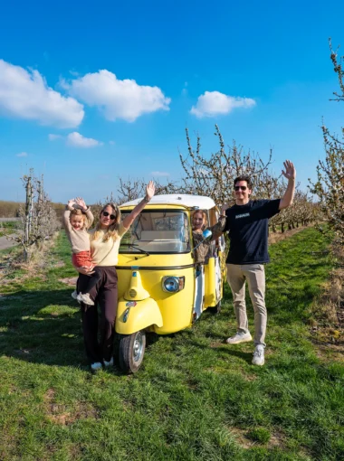 Gezin bij een gele Ape Calessino tijdens de Bambinotour tussen fruitboomgaarden in Tongeren-Borgloon, Belgisch Limburg.
